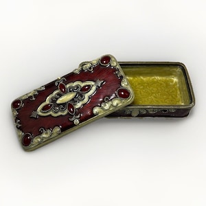 May include: A rectangular metal trinket box with a hinged lid. The lid features a deep red enamel finish with gold-tone filigree accents and inset red cabochon stones. The interior of the box is lined with a textured, pale yellow enamel finish.