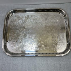 Puede incluir: Una bandeja rectangular plateada con intrincados diseños florales y de volutas. La bandeja tiene un borde elevado con un borde decorativo. La superficie es reflectante, y el diseño es clásico y elegante.