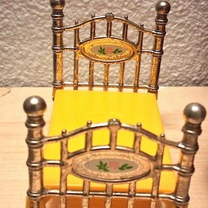 May include: A miniature gold-colored bed with a yellow mattress. The headboard and footboard feature an oval design with a floral motif. The bed frame has a detailed, ornate design, perfect for a dollhouse or miniature collection.