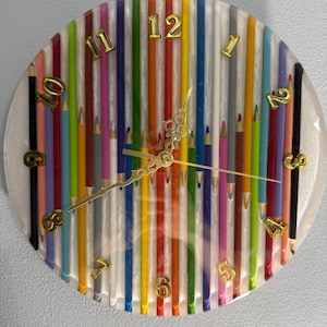 Puede incluir: Reloj de pared redondo con un diseño único. La esfera del reloj presenta una colorida variedad de lápices verticales, con números dorados que marcan las horas. Las manecillas del reloj también son doradas, creando un reloj vibrante y artístico.