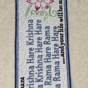 May include: A rectangular fabric bookmark with a white background featuring a light gray swirling pattern. It is embroidered with a pink lotus flower, green vines, and blue text that reads: Please chant Hare Krishna Hare Krishna Krishna Krishna Hare Hare Hare Rama Hare Rama Rama Rama Hare Hare ...and your life will be sublime.