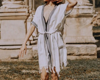 May include: A woman wearing a white cotton robe with black embroidery and fringe details. The robe is tied at the waist with a black belt. She is standing in front of a stone structure.