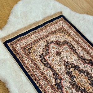 Puede incluir: Una alfombra de oración tradicional con intrincados patrones florales en tonos dorados, rojos y blancos sobre un fondo negro. La alfombra tiene un borde con flecos.