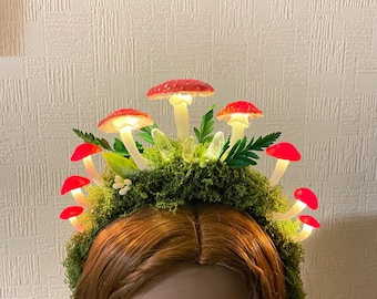 Könnte beinhalten: Ein rotes und weißes Pilz-Haarband mit leuchtenden Lichtern. Das Haarband ist mit grünem Moos und Blättern verziert.