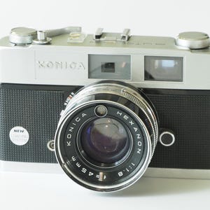 May include: A silver and black Konica rangefinder camera featuring a Konica Hexanon 45mm f/1.8 lens. The camera body has a textured black grip, a silver top plate with a film advance lever, and a small circular sticker that reads NEW LOADING SYSTEM.