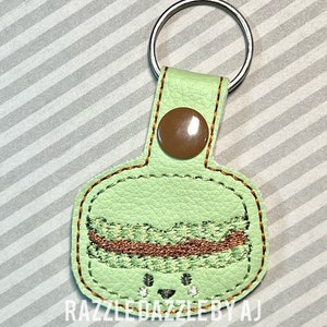 May include: A green leather keychain with a brown button and a stitched design of a green and brown layered dessert with a smiling face. The keychain has the text "Razzle Dazzle by AJ" at the bottom.