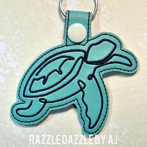 May include: A teal blue leather keychain with a black outline of a turtle. The keychain has a silver key ring and a white button.