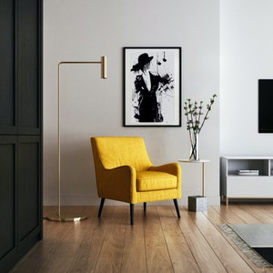 Puede incluir: Un salón moderno con un sillón amarillo, una lámpara de pie dorada y una impresión artística en blanco y negro enmarcada. La habitación tiene pisos de madera y una pared blanca. Una pequeña mesa auxiliar sostiene un jarrón con flores.