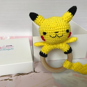 May include: A yellow crocheted Pikachu character with black ears and a red blush on its cheeks is attached to a wooden teething ring. The toy is packaged in a white box with the text "Yarn Tangle" printed on it.