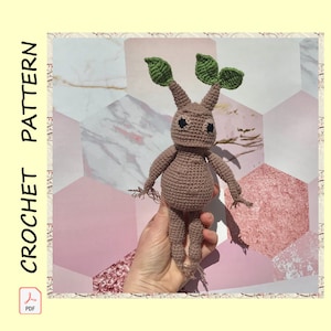 Pode incluir: Um brinquedo amigurumi de crochê em forma de mandrágora marrom com folhas verdes. O brinquedo está sendo segurado em uma mão.