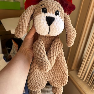 May include: A crocheted brown and white dog plush toy with black eyes and a black nose. The toy is being held by a hand.
