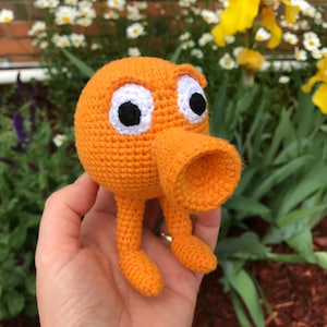 Puede incluir: Un juguete de peluche naranja tejido a crochet con ojos blancos y un gran hocico naranja. El juguete tiene dos patas cortas y se sostiene en una mano.