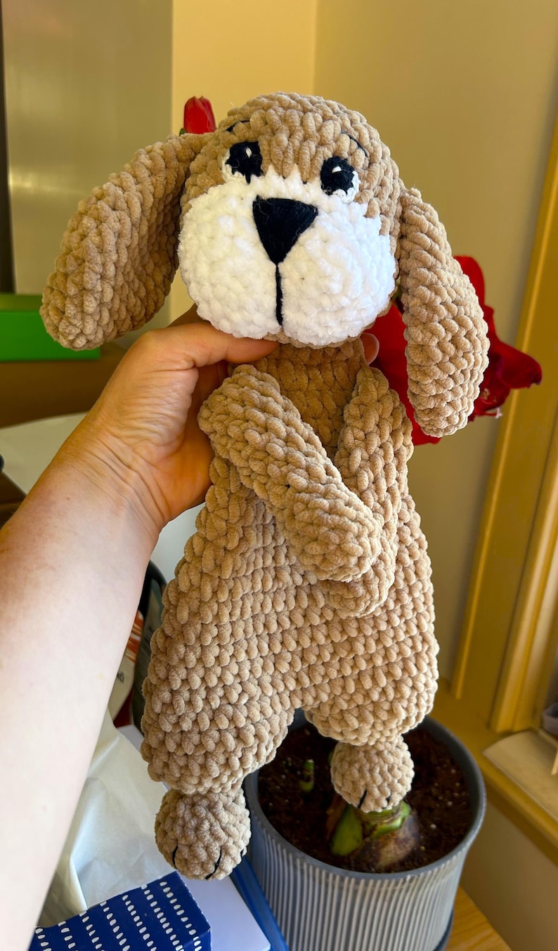 May include: A crocheted brown and white dog plush toy with black eyes and a black nose. The toy is held by a hand in front of a potted plant.