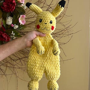 May include: A handmade yellow plush toy resembling Pikachu, featuring black-tipped ears, red cheek circles, and a smiling mouth. The plush is made of a soft, textured material and is held up against a neutral background.