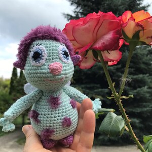 May include: A crocheted plush toy with a light blue body, purple hair, and pink nose. The toy has large blue eyes with white pupils and a smiling mouth. The toy is being held in a hand in front of a pink rose.