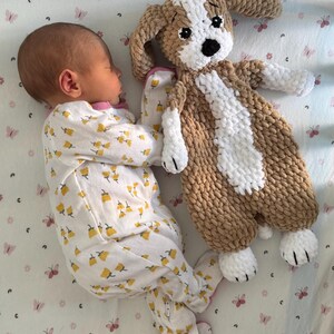 May include: A baby wearing a white and yellow floral sleepsuit is sleeping with a brown and white crocheted dog toy.