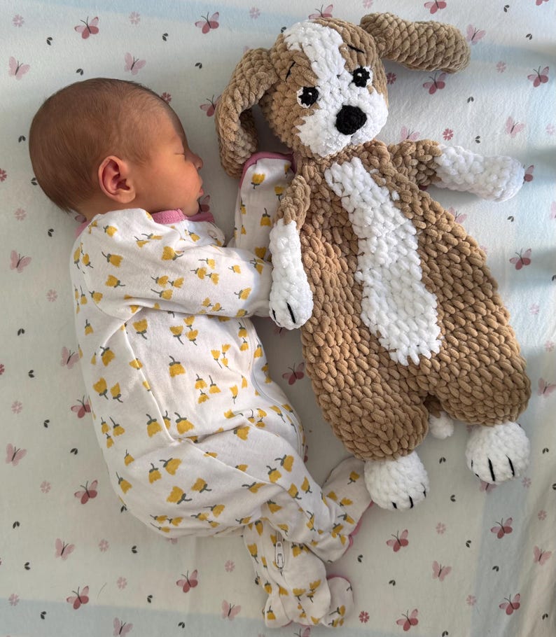 May include: A baby wearing a white and yellow floral sleepsuit is sleeping with a brown and white crocheted dog toy.