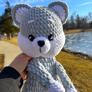 以下が含まれることがあります： グレーと白の毛糸で編まれたクマの形をしたぬいぐるみ。クマは白い顔に黒い目と黒い鼻をしています。