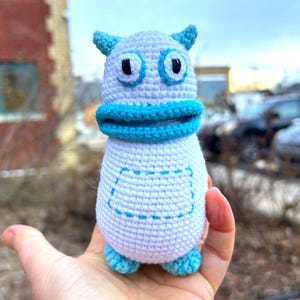 May include: A handmade crochet monster toy in shades of light purple and turquoise. The toy has large black eyes, a turquoise mouth, and small horns. The body features a stitched pocket design and turquoise feet.