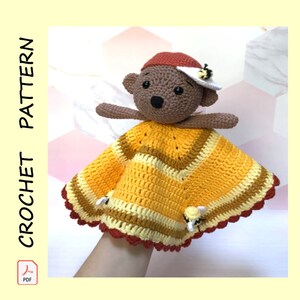 May include: A crocheted honeybee-themed baby blanket with a brown bear attached. The blanket is yellow, orange, and red with a scalloped edge. The bear is wearing a red hat with a white bee on top.