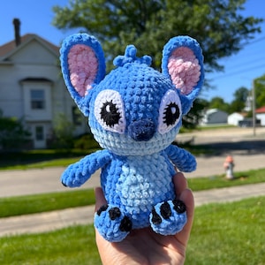 May include: A blue crochet plush toy of a cartoon character with large ears, black eyes, and a dark blue nose. The plush has a light blue belly and black accents on its feet. The ears have pink inner linings.