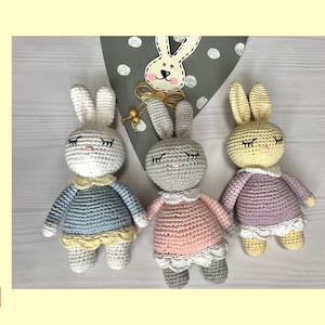 May include: Three crocheted bunny amigurumi toys. The bunnies are wearing different coloured dresses: blue, pink, and purple. The bunnies have closed eyes and are sitting on a light wood surface.