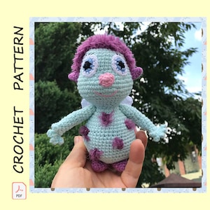 May include: A crocheted doll with a light blue body, purple hair, and accents. The doll has a pink nose, blue eyes, and small white wings. The image includes the text "CROCHET PATTERN" and a PDF icon.