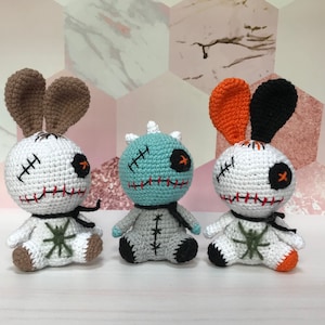 May include: Three crocheted plush toys in the shape of rabbits. The rabbits are white with black eyes and red stitching. One rabbit has brown ears, one has orange ears, and one has black ears. The toys are sitting on a white surface.