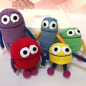 May include: Five crocheted plush toys in different colors, including purple, blue, green, red, and yellow. Each toy has a smiling face with two large eyes and a small mouth. The toys are all standing on a white surface.