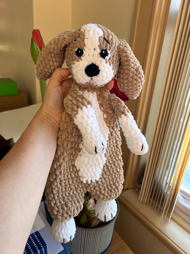 May include: A crocheted plush toy dog with brown and white fur. The dog has black eyes and a black nose. The dog is holding its paws up in the air.