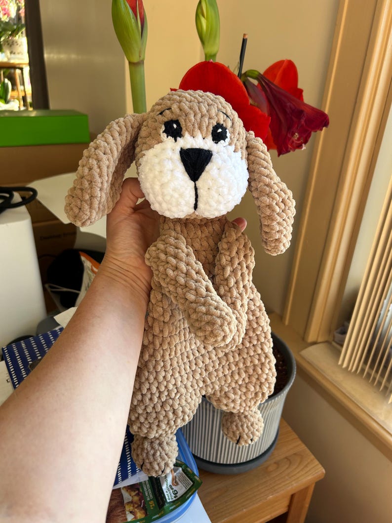 May include: A crocheted plush toy dog with brown fur, white face, and black eyes. The dog is being held by a hand.