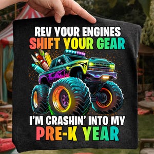 May include: Black graphic tee with a monster truck design. The truck is teal, purple, and yellow with large teal tires. Text reads "REV YOUR ENGINES SHIFT YOUR GEAR I'M CRASHIN' INTO MY PRE-K YEAR."