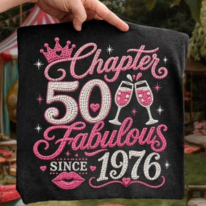 May include: A black t-shirt featuring embroidered text and graphics in pink and white. The design includes a crown, two champagne glasses, a pair of lips, and the text: Chapter 50 Fabulous SINCE 1976, accented with small star and heart shapes.