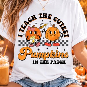 May include: A white t-shirt featuring a graphic of two cartoon pumpkins with arms and legs. One pumpkin wears a pink bow and holds a book, while the other holds a pencil. The text reads I TEACH THE CUTEST PUMPKINS IN THE PATCH with a checkerboard.