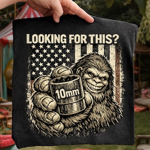 Puede incluir: Camiseta negra con un gráfico de un Bigfoot sonriente sosteniendo una llave de vaso de 10 mm frente a una bandera estadounidense desgastada. El texto "LOOKING FOR THIS?" está impreso encima del gráfico.