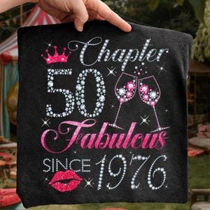 May include: A black fabric square featuring a graphic print with the text Chapter 50 Fabulous SINCE 1976. The design includes a pink crown, two champagne glasses filled with pink gems, and a pair of pink lips, all rendered in a sparkling diamond effect.
