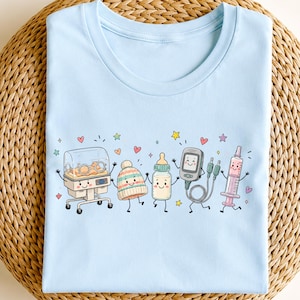 May include: A light blue t-shirt featuring a graphic print of five anthropomorphic medical items: a neonatal incubator, a striped knit hat, a baby bottle, a medical monitor with cables, and a syringe, all surrounded by small colorful stars and hearts.