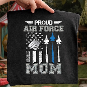 May include: Black Air Force Mom t-shirt with a patriotic design. The shirt features the text "PROUD AIR FORCE MOM" in white and camouflage lettering. Includes an American flag, dog tag, and jet illustrations.