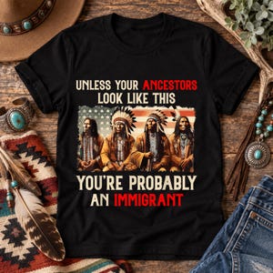 May include: A black short-sleeved t-shirt featuring a graphic of four Indigenous people in traditional attire against a faded American flag. The text reads: UNLESS YOUR ANCESTORS LOOK LIKE THIS YOU'RE PROBABLY AN IMMIGRANT, with specific words in red.