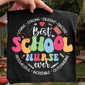 Könnte beinhalten: Schwarzes quadratisches Kissen mit dem Schriftzug "Best School Nurse Ever" in bunten Buchstaben. Das Design beinhaltet ein Stethoskop, Pillen und die Worte "Loving, Strong, Diligent, Sincere, Resourceful, Incredible, Outstanding."