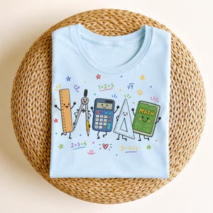 May include: A light blue t-shirt featuring a colorful graphic of cartoon school supplies including a ruler, compass, calculator, triangle, and a green book labeled MATH, surrounded by small math equations and stars.
