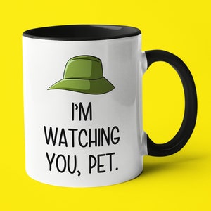 May include: White ceramic mug with a black rim and handle. The mug features a cartoon illustration of a green hat with the text "I'M WATCHING YOU, PET."