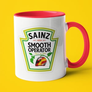 Puede incluir: Taza de cerámica blanca con borde y asa rojos. La taza presenta una ilustración de estilo de dibujos animados de un casco de piloto de carreras de coches con el texto "Sainz Est. 1994 Est. Smooth Operator SS is the number".
