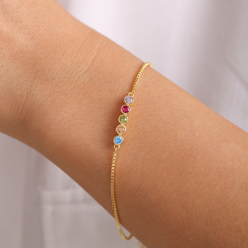 Puede incluir: Un brazalete de cadena dorada con una fila de cinco piedras preciosas de colores. Las piedras preciosas son azul claro, melocot&oacute;n, verde, rojo y morado.