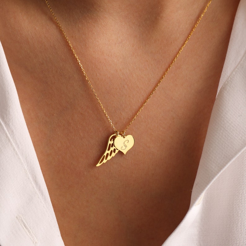 Pu&ograve; includere: Una collana dorata con una catena sottile. Il ciondolo presenta un ciondolo a forma di ala e un ciondolo a forma di cuore con la lettera "S" incisa. La collana &egrave; indossata su una camicia bianca.