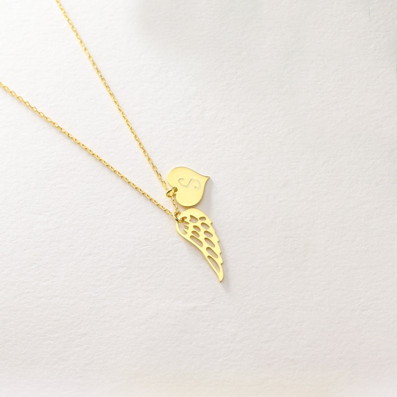 Pu&ograve; includere: Una collana color oro con una catena sottile. Il ciondolo presenta un cuore con la lettera "S" incisa e un ciondolo a forma di ala stilizzata. La collana &egrave; esposta su uno sfondo bianco.