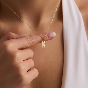 May include: A gold-colored necklace with a rectangular pendant. The pendant has the letter "M" engraved on it, along with a small, heart-shaped gemstone. The necklace is delicate and appears to be made of a fine chain.