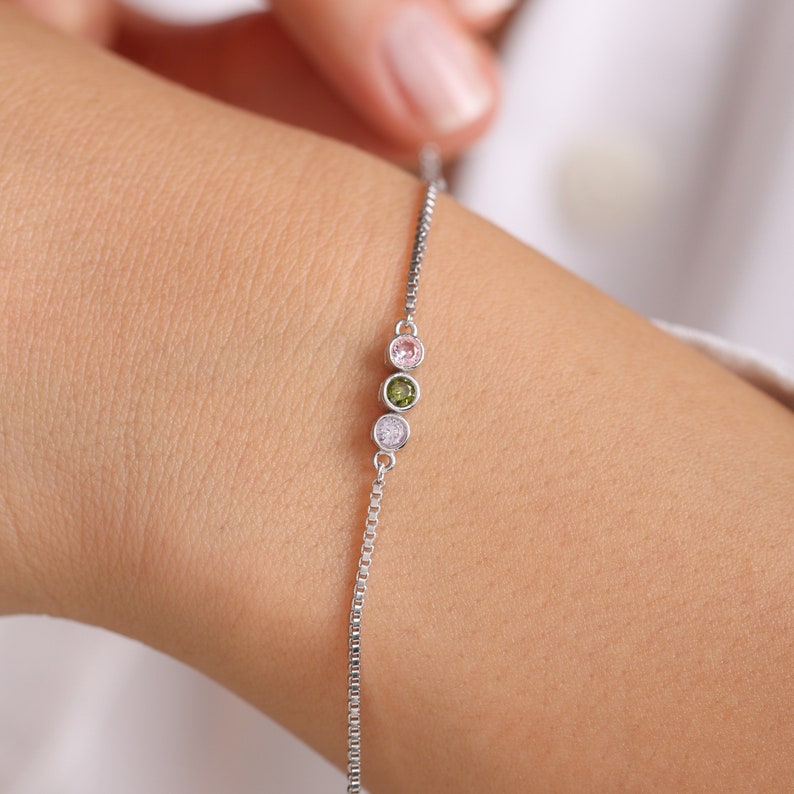 Puede incluir: Un brazalete de cadena de plata con tres peque&ntilde;as piedras redondas. Las piedras son de color rosa, verde y transparente.