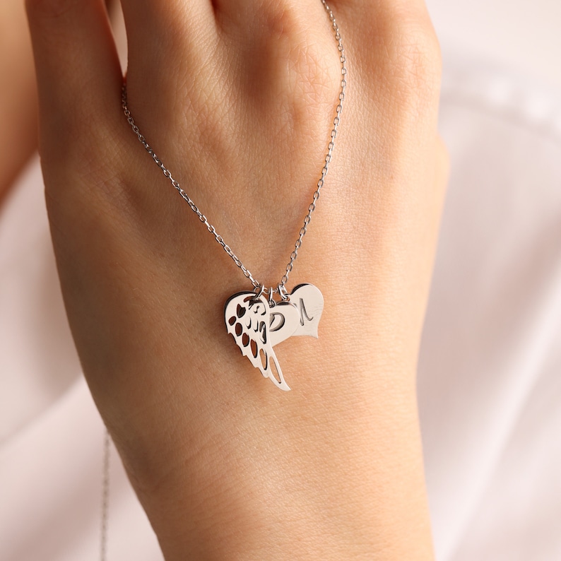 Pu&ograve; includere: Una collana d'argento con tre ciondoli. Un ciondolo &egrave; un cuore, un altro &egrave; un'ala e il terzo &egrave; un cuore con la lettera "M" incisa. La collana &egrave; mostrata su uno sfondo morbido e neutro.