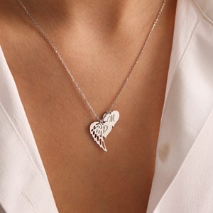 Pu&ograve; includere: Collana d'argento con ciondolo a forma di cuore e ala. Il ciondolo presenta due ciondoli a forma di cuore, uno con un design ad ala e l'altro con le iniziali "M" e "P". La collana &egrave; indossata su una camicia bianca.